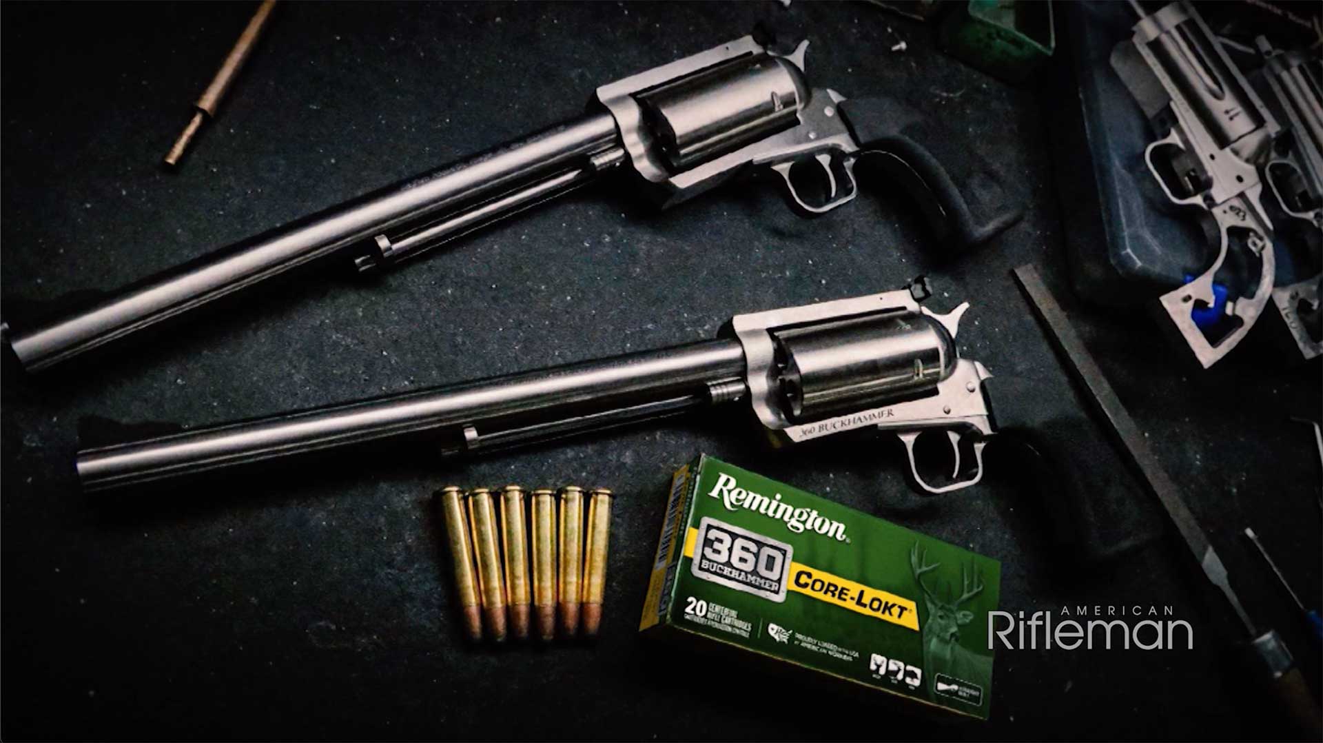 Two Magnum Research BFR revolvers lying on a black table next to a box of Remington 360 Buckhammer and six loose brass-colored cartridges.