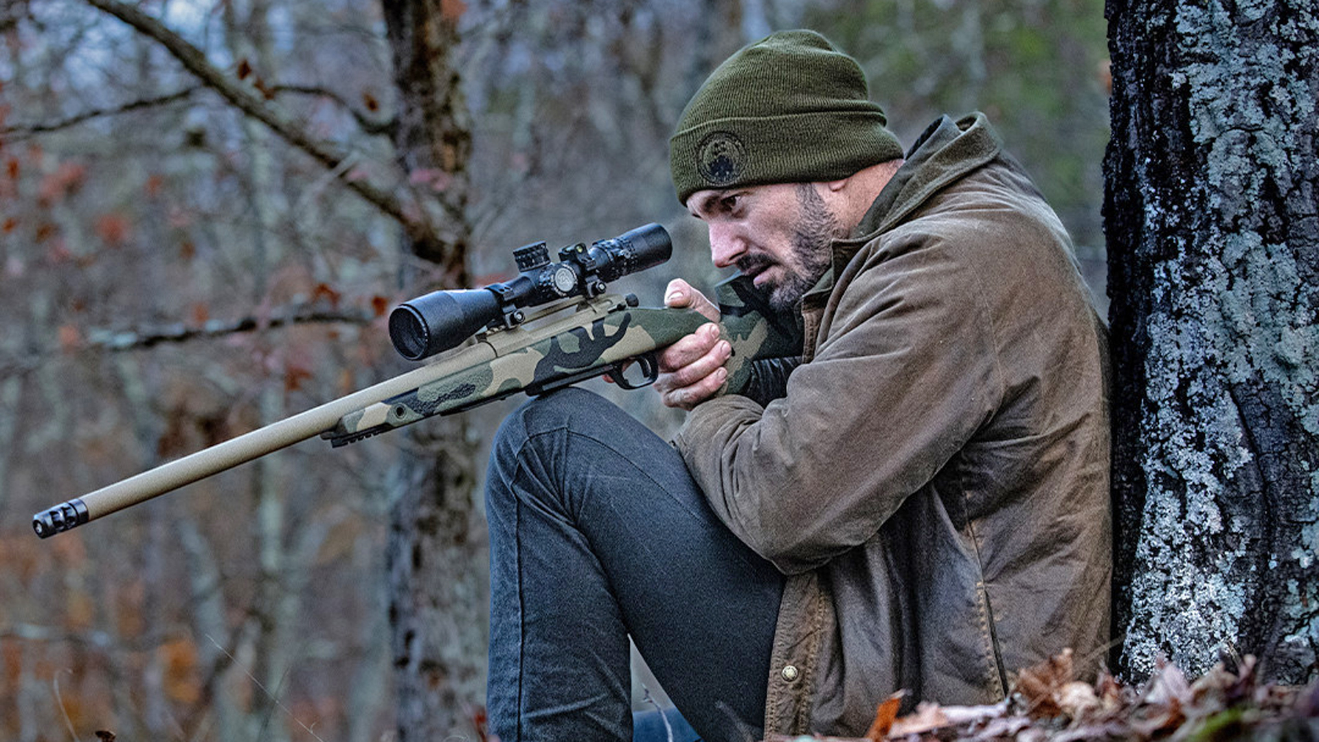 Man leaning against a tree while aiming the Watchtower Bridger bolt-action rifle.