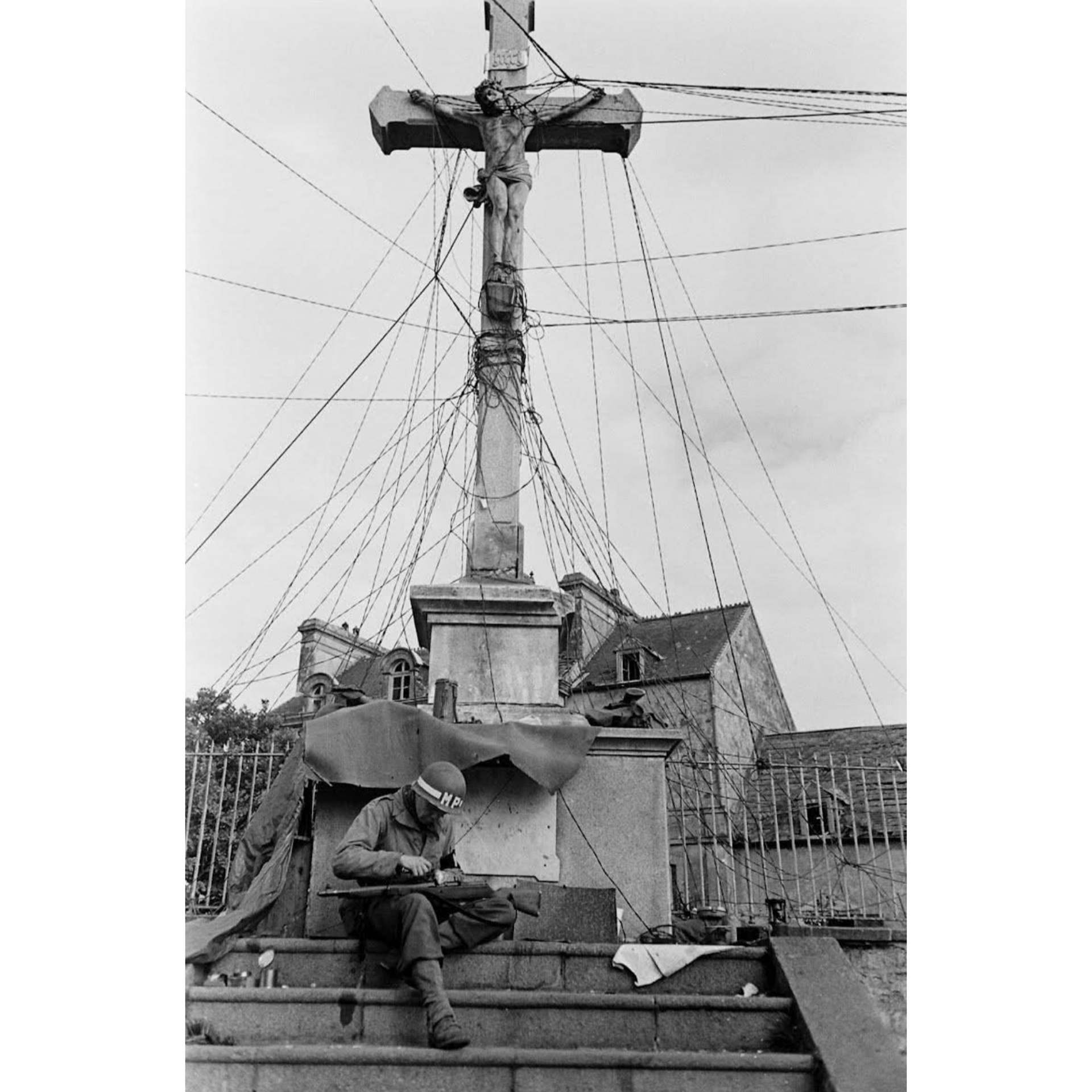 A military policeman carefully cleans his Gew. 41 semi-automatic rifle on steps beneath stone concrete crucifix