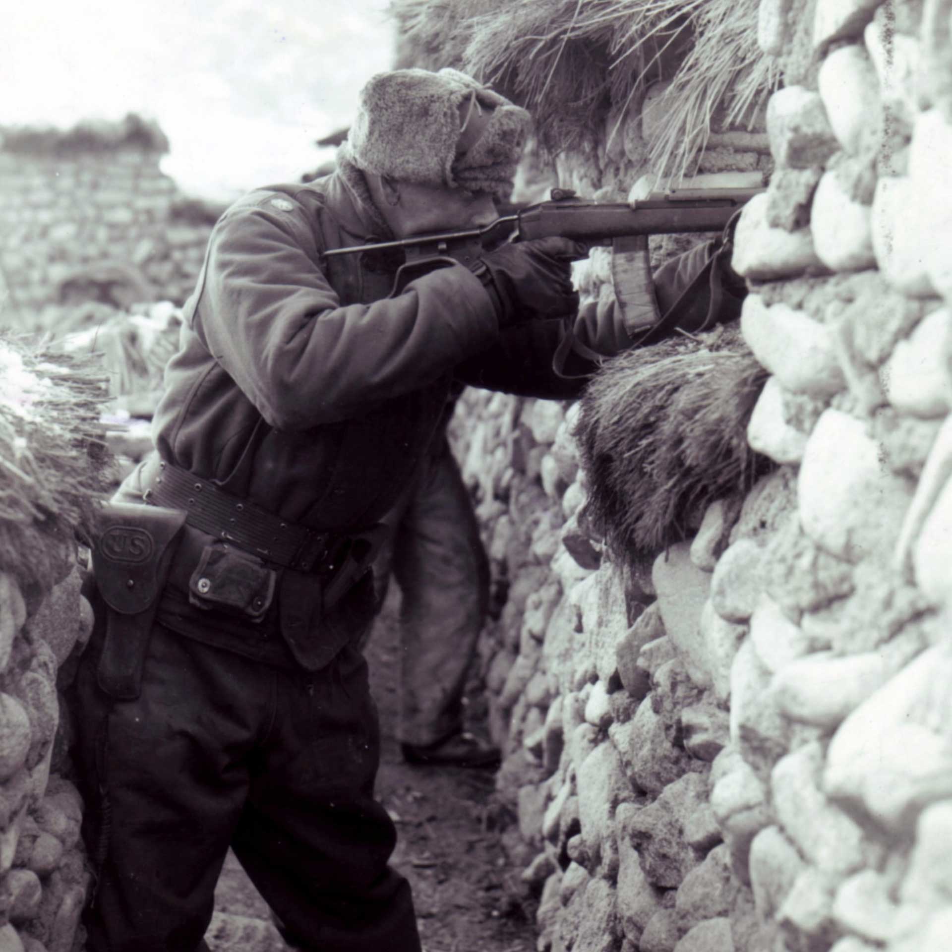 A U.S. Army soldier aiming the M2 carbine behind a rock wall in Korea.