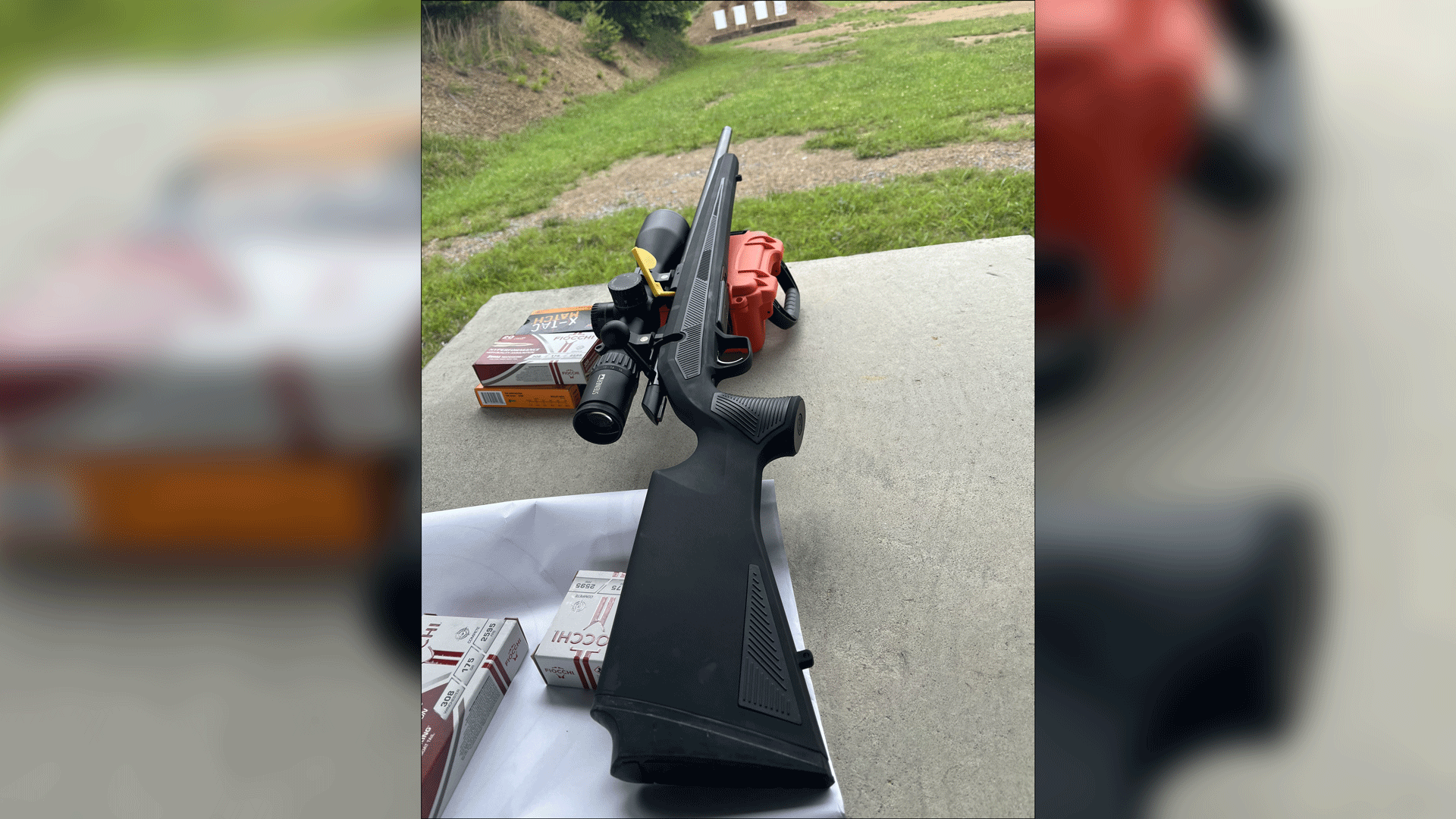 CZ 600 bolt-action rifle on shooting bench pointed downrange with yellow chamber flag and shown with ammunition boxes orange case and black riflescope attached
