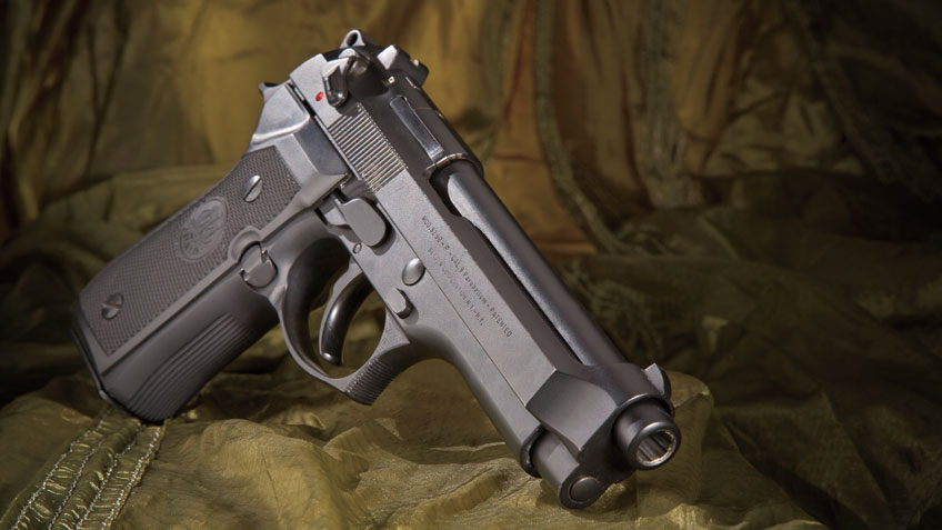 An all-black Beretta M9 propped up against a green, military-like background.