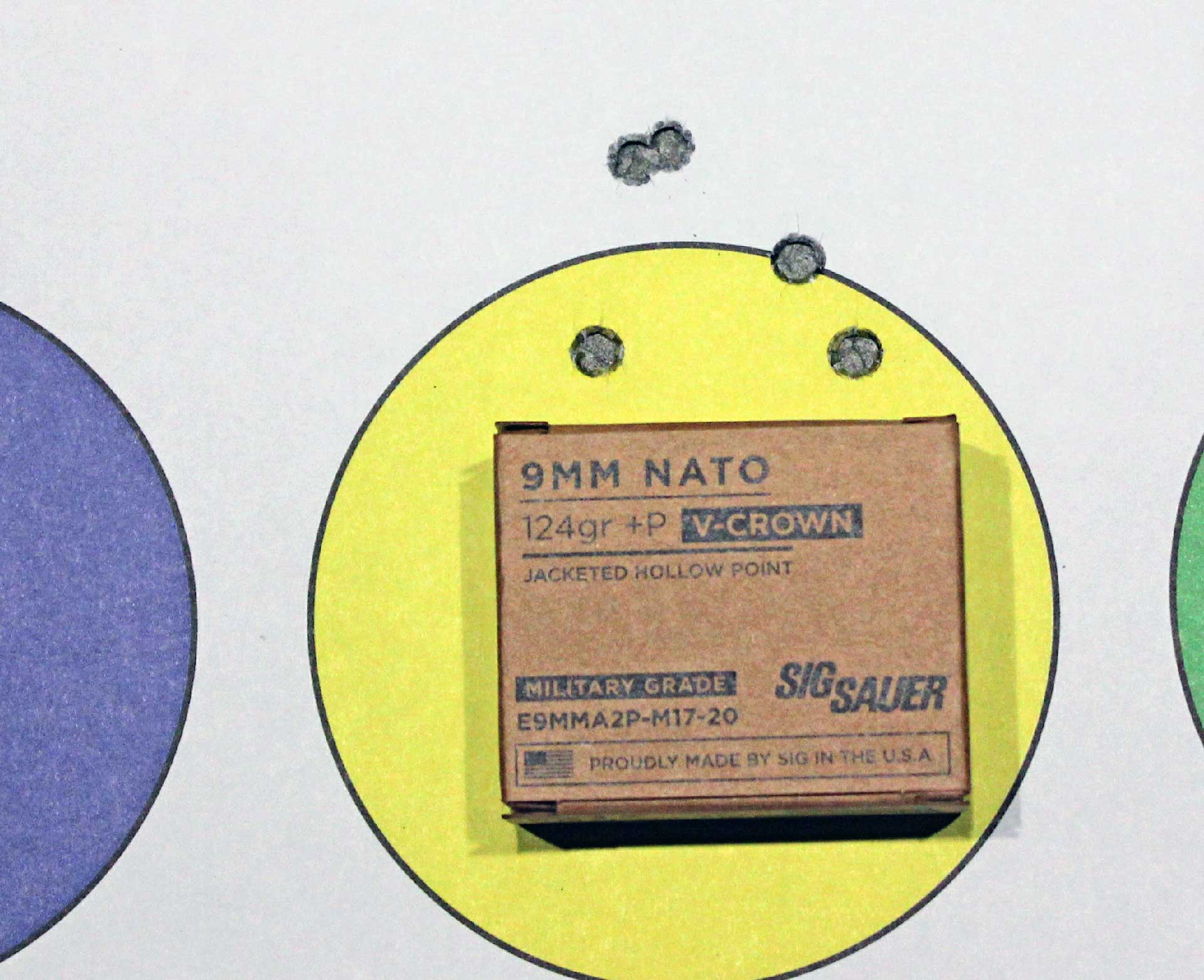Bullet holes in a yellow circular target above a box of SIG Sauer M17 V-Crown 9 mm Luger ammunition.