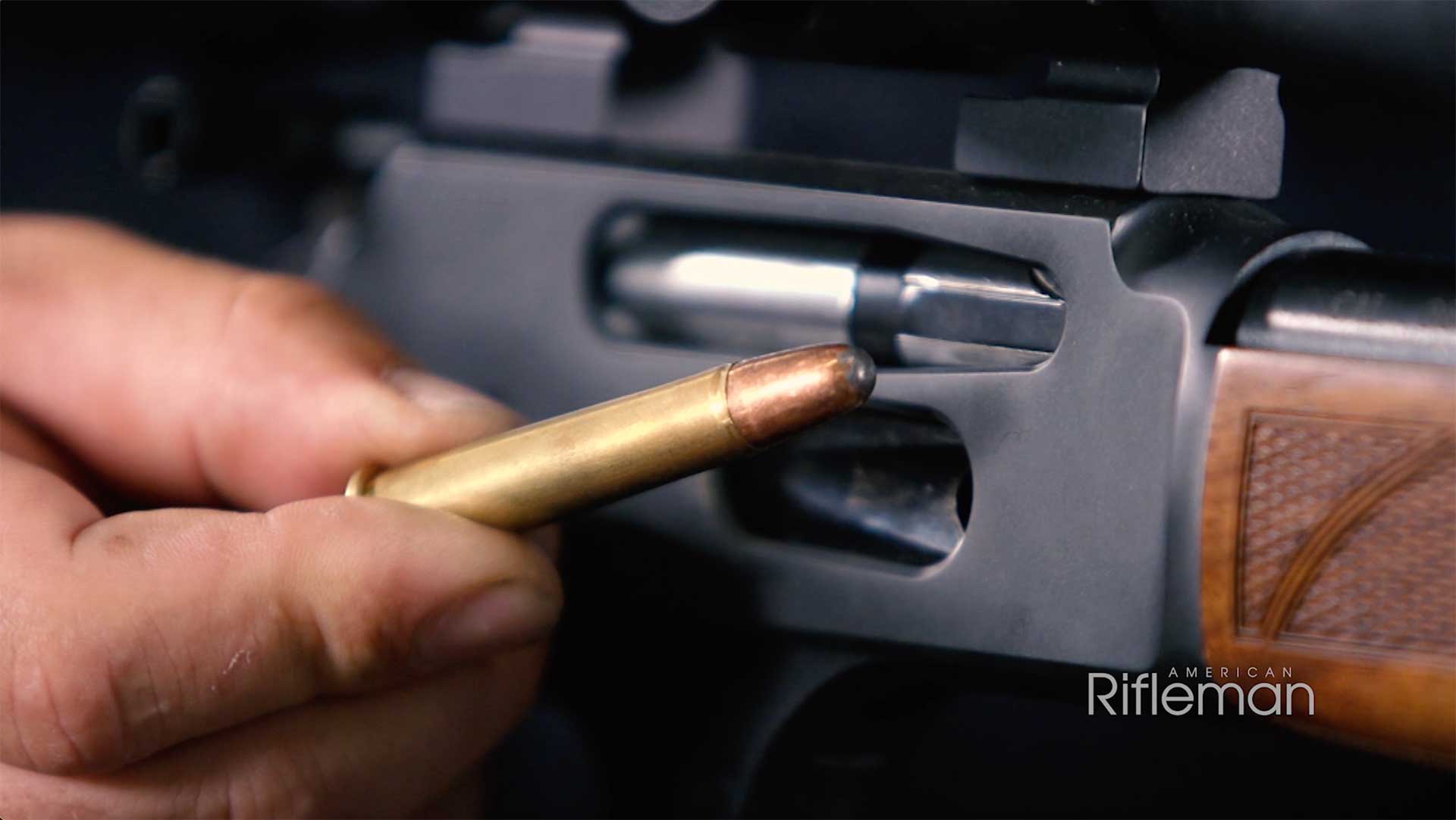 A man holds a Remington 360 Buckhammer cartridge next to the black receiver of a Henry lever-action rifle.