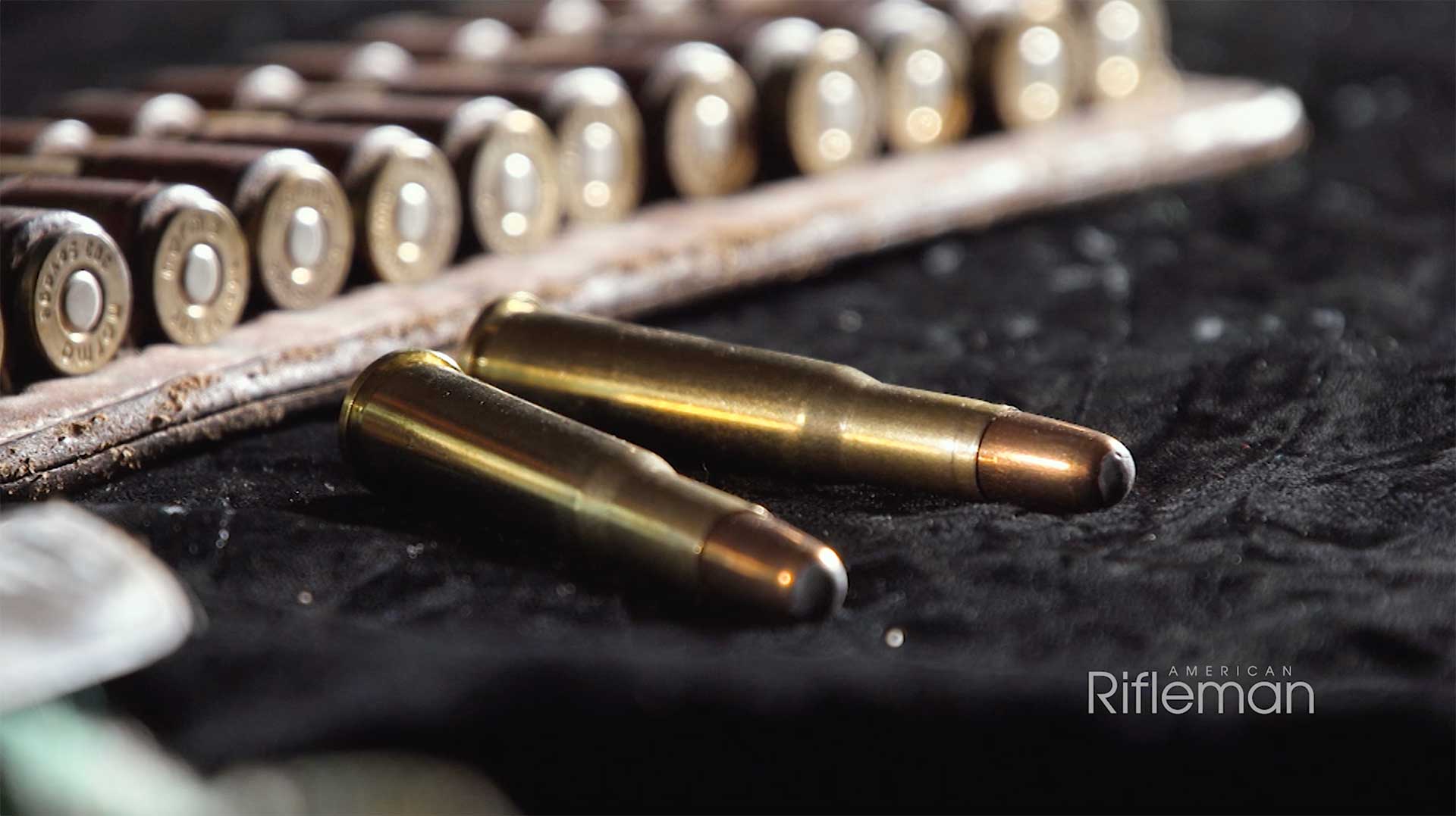 Two .303 Savage cartridges lying on a black cloth with a row of cartridge cases behind.