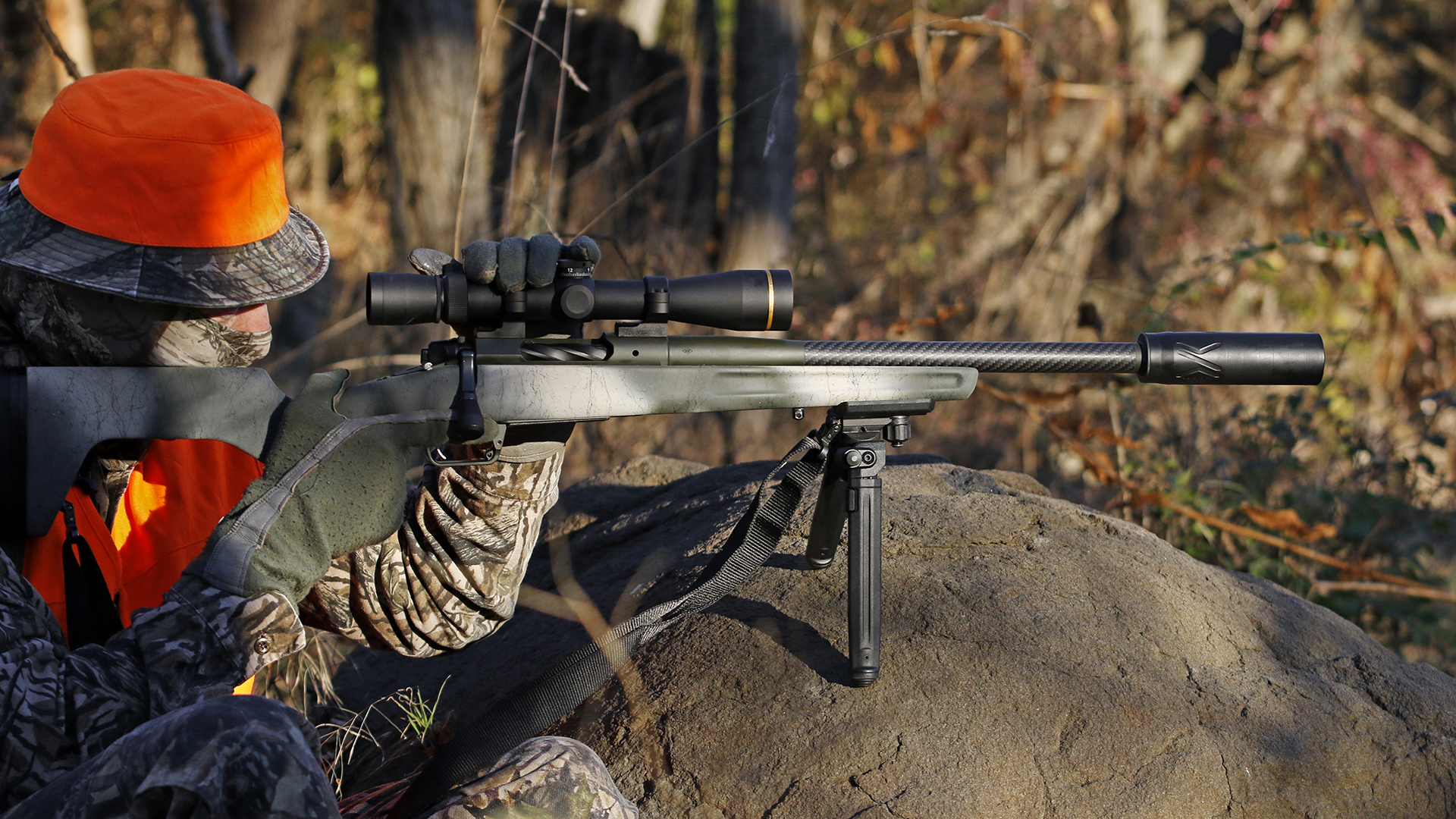 Camouflaged hunter with blaze orange safety colors perched on a rock with bolt-action springfield armory redline rifle carbon-fiber barrel suppressor outdoors