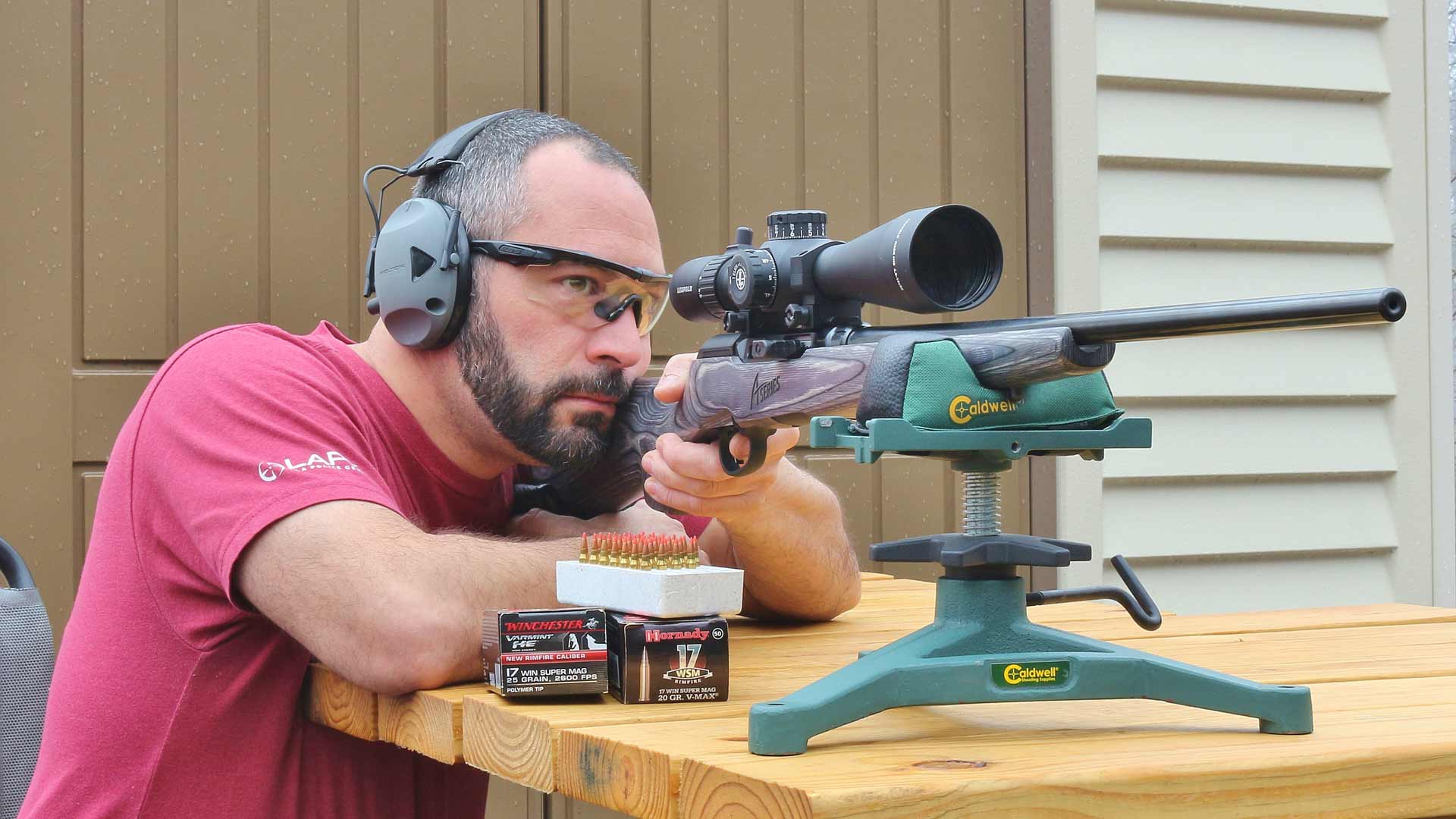 Bearded man wearing earmuffs and protective glasses sitting at wood table behind savage a17 semi-automatic rifle