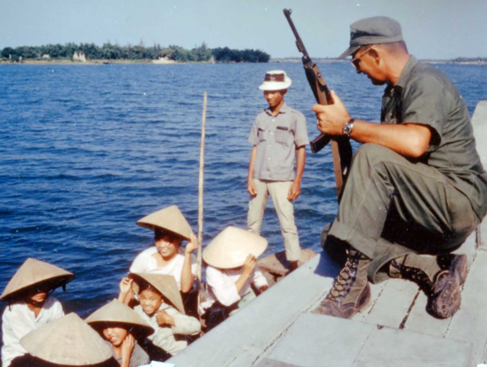 A U.S. soldier overlooks a boat of Vietnamese civilians while armed with an M1 carbine.