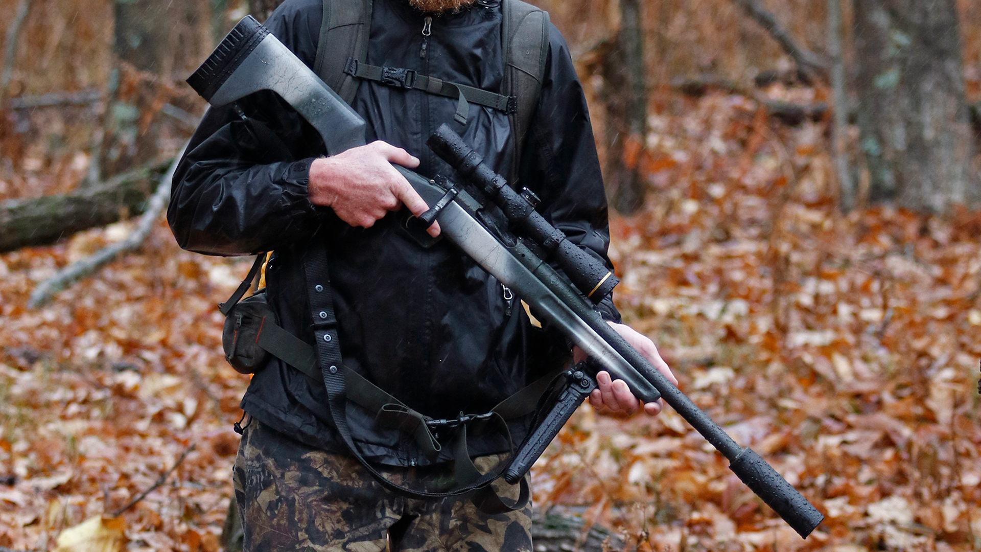 Hunter with Springfield Armory Redline bolt-action hunting rifle held across the body two hands holding rifle pointed at ground with silencer central suppressor leupold riflescope raining wet woods finger off trigger bipod deployed