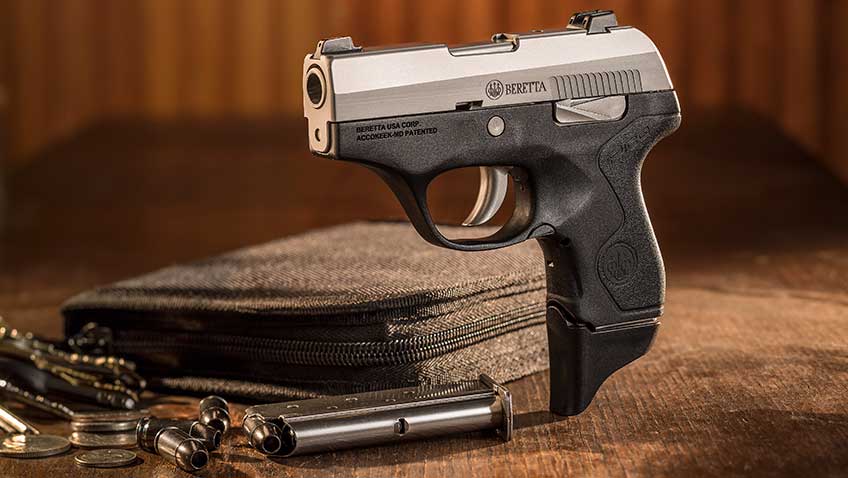 A Beretta Pico standing up on a wooden table, with ammunition, a loaded magazine and a black gun case in the background.