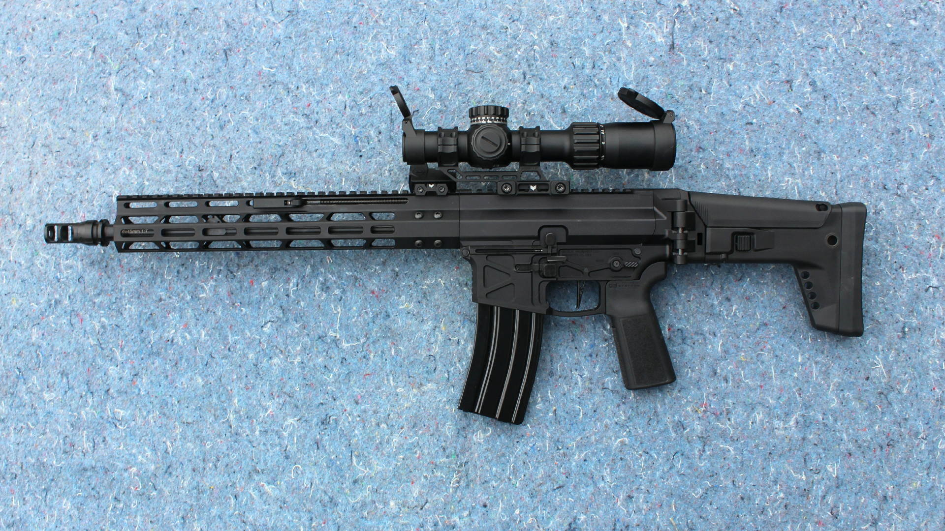 Review: Global Ordnance Monolith Bufferless rifle left-side view full-length black gun shown with riflescope on blue background