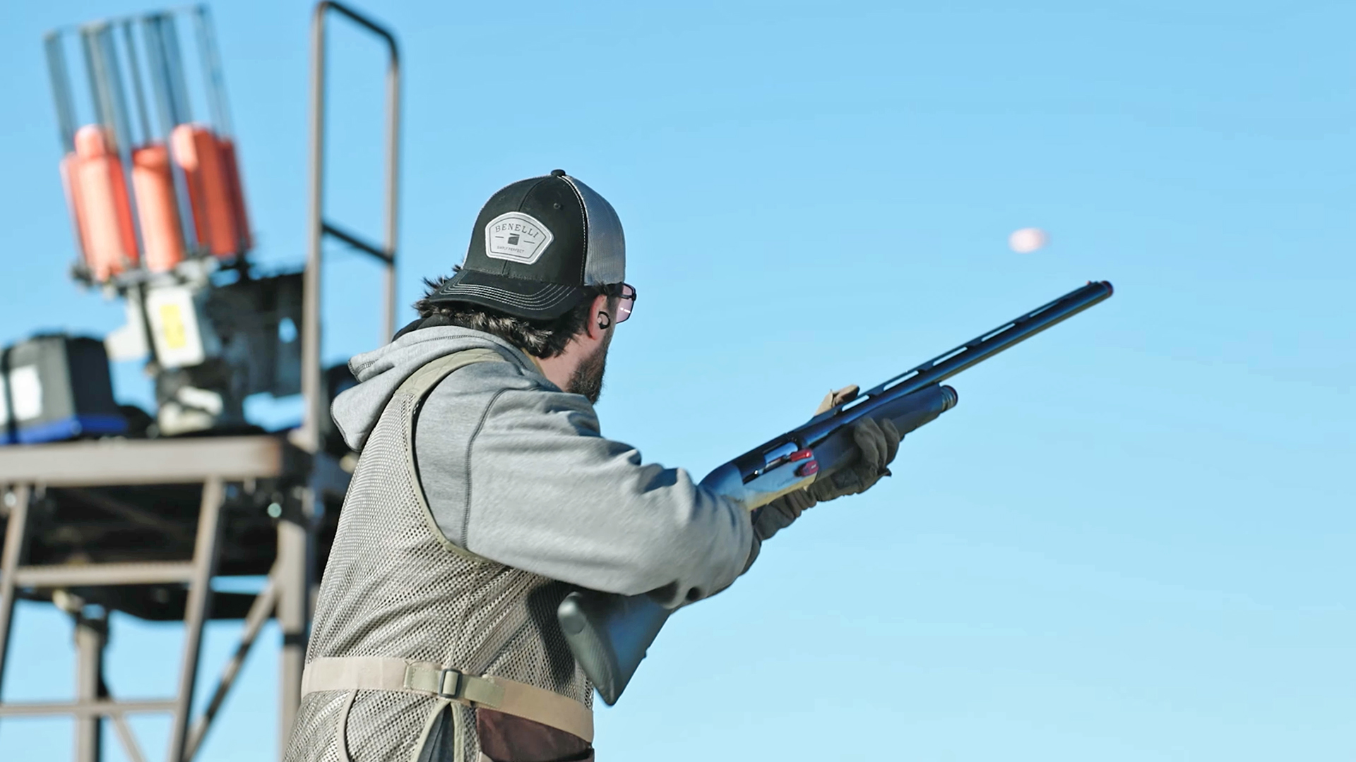 Sporting clays shooter aiming a Benelli shotgun toward an orange clay pigeon in flight.