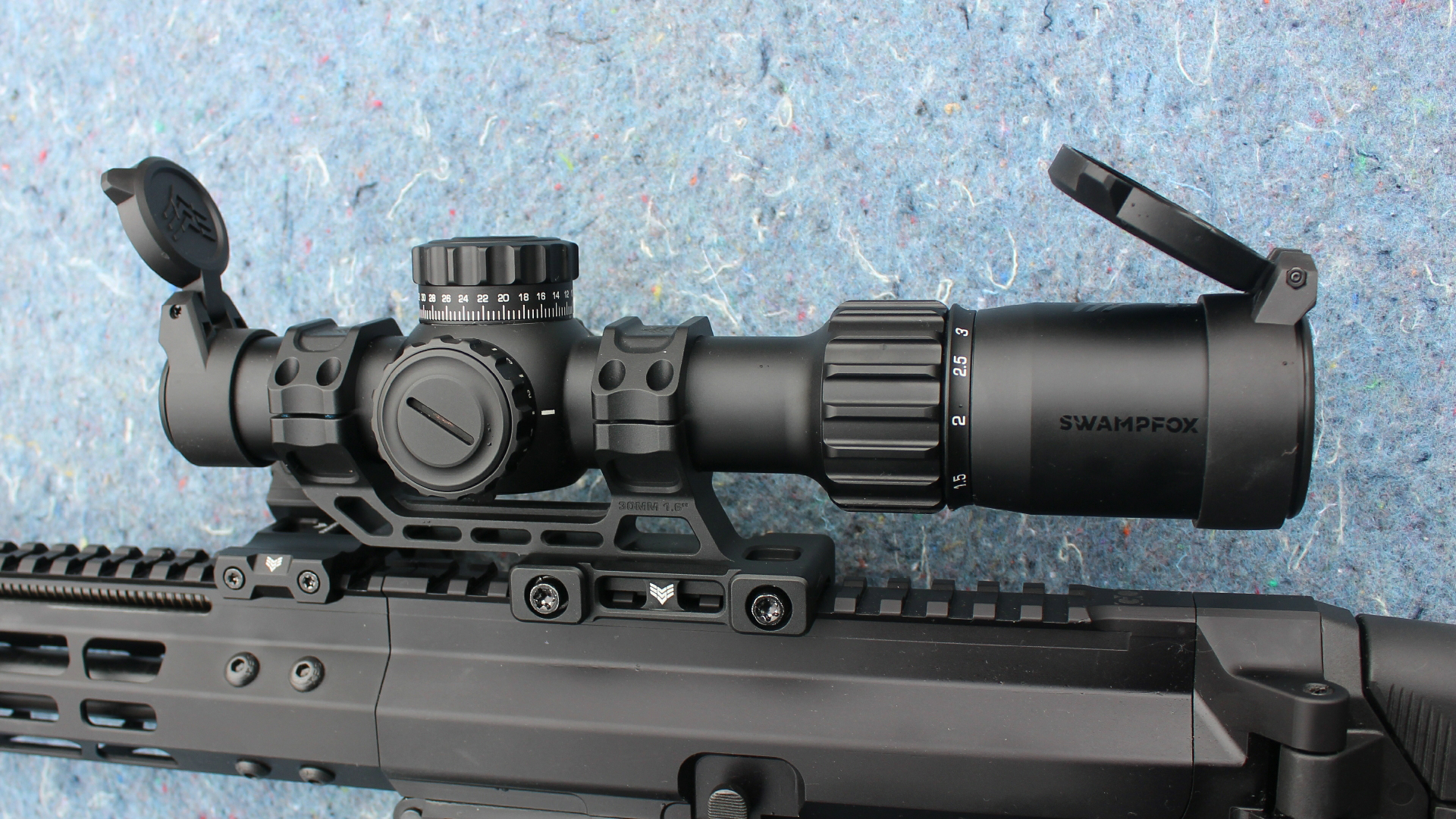 Review: Global Ordnance Monolith Bufferless rifle closeup receiver top with Swamp Fox Tomahawk II low poer variable optic with flip-up covers shown on blue background