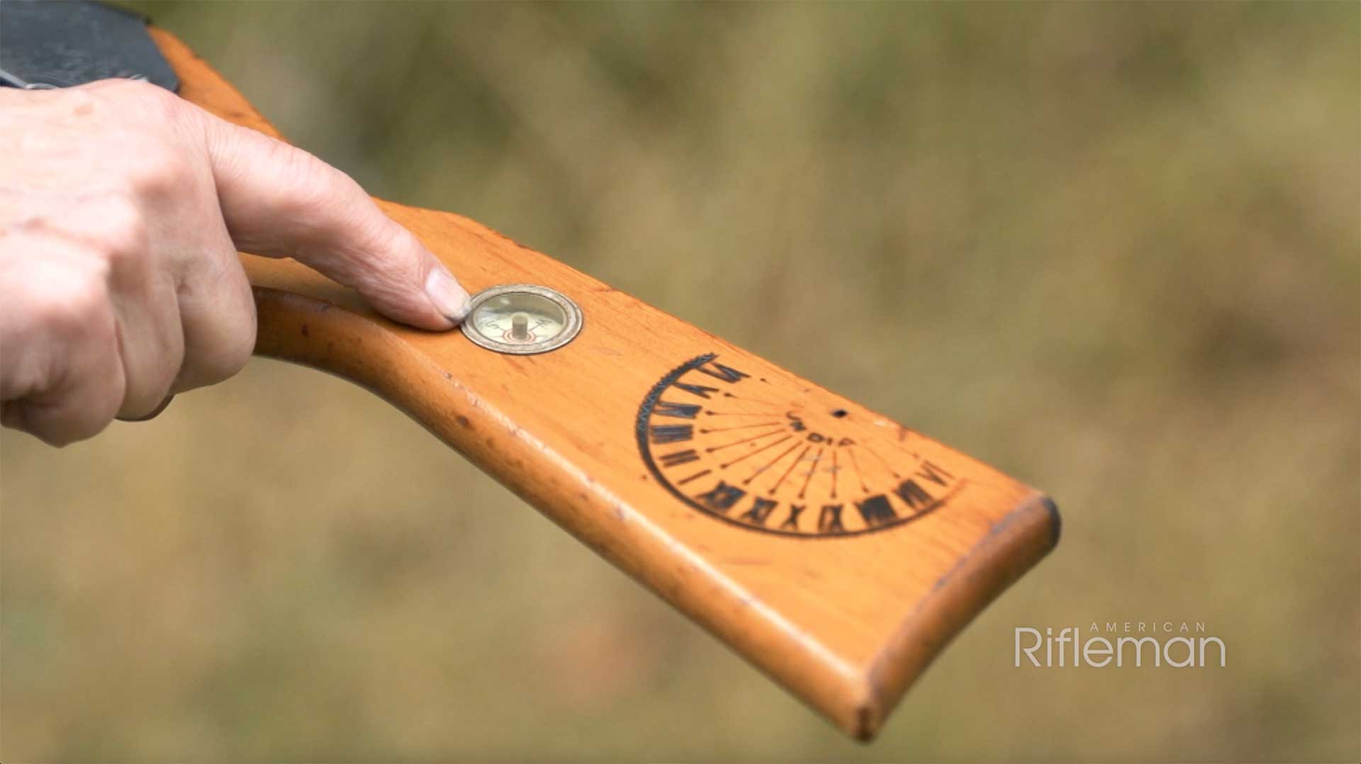 A man's finger pointing out the compass and sundial on the buttstock of a Daisy Buck Jones BB gun.