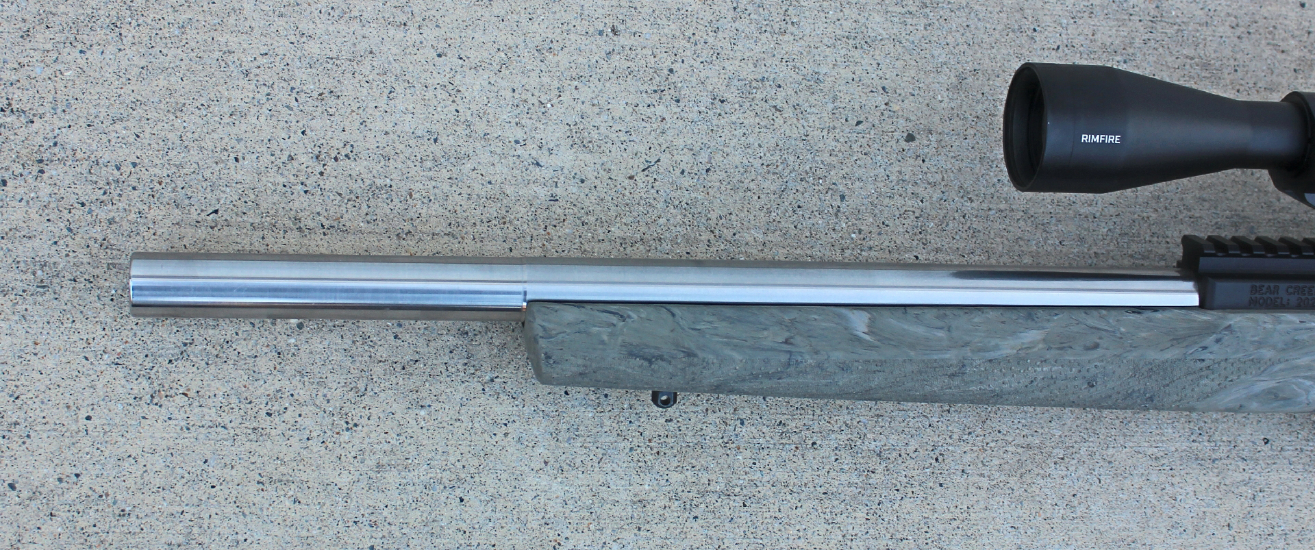 Closeup view of Bear Creek Arsenal BC-202 rimfire rifle stainless steel silver-colored bull barrel shown with brown camouflage rile stock and black riflescope and receiver partly showing on right side