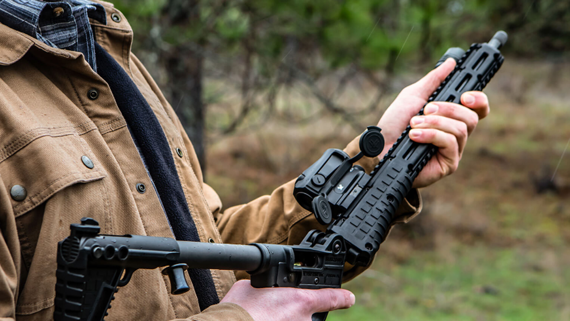 KelTec SUB2000 GEN3 folding pistol-caliber carbine shown outdoors in the hand unlocking the folding mechanism grass trees soft focus background man watching.